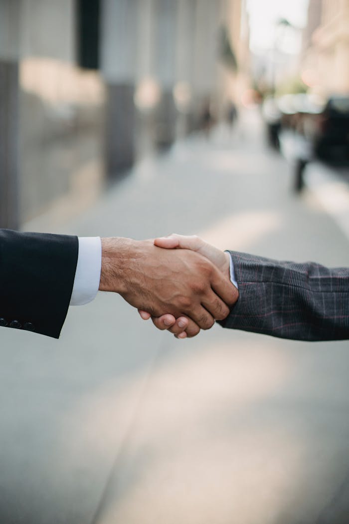 Accueil Two businessmen shaking hands outside, symbolizing cooperation and agreement in a city setting.